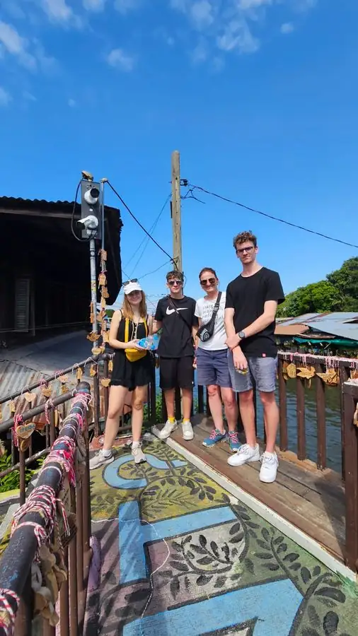 A family of four posing together on a canal-side wooden walkway painted with a colorful mural, strings of wooden wish charms hanging along the railing
