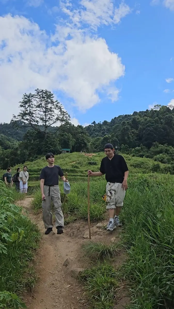 Two hikers walking a grassy hillside trail with walking sticks, green forested hills and blue sky behind them