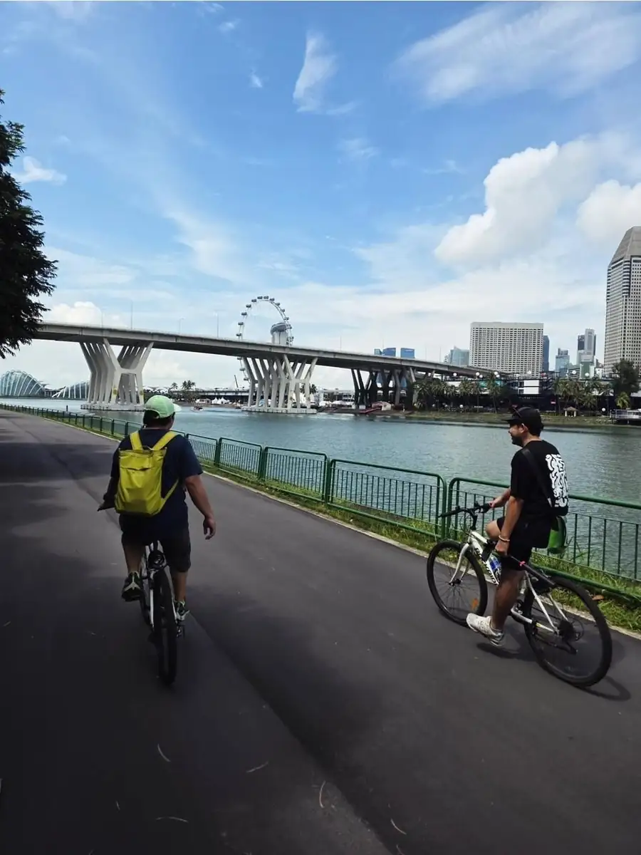 Two cyclists riding along a waterside path in Singapore, with the Singapore Flyer and a bridge in the background